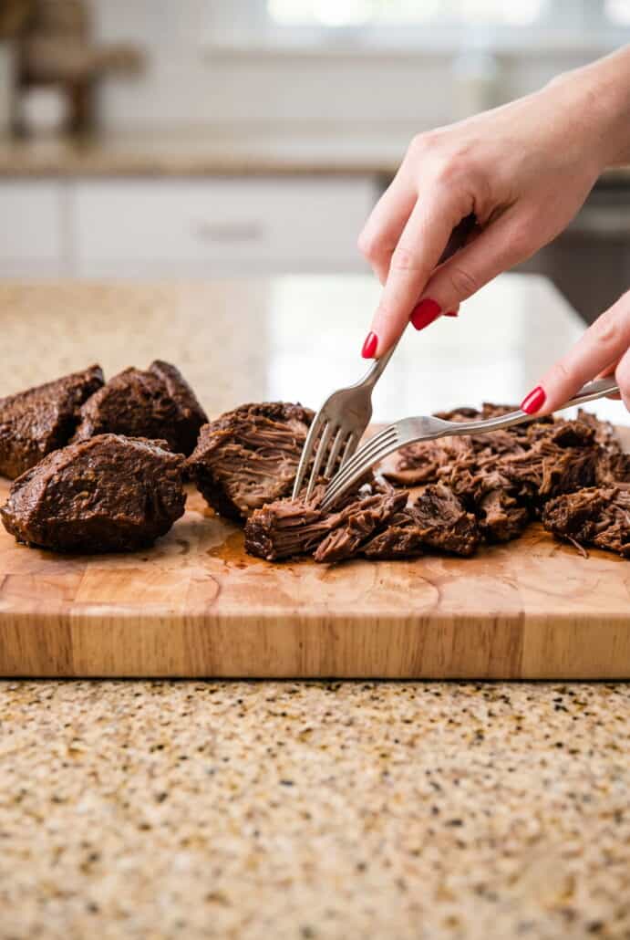 Tender crockpot birria beef being shredded with forks for serving in consommé or tacos.