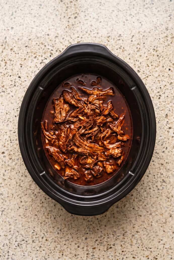 Shredded Mexican birria beef simmering in rich red consommé inside a crockpot.