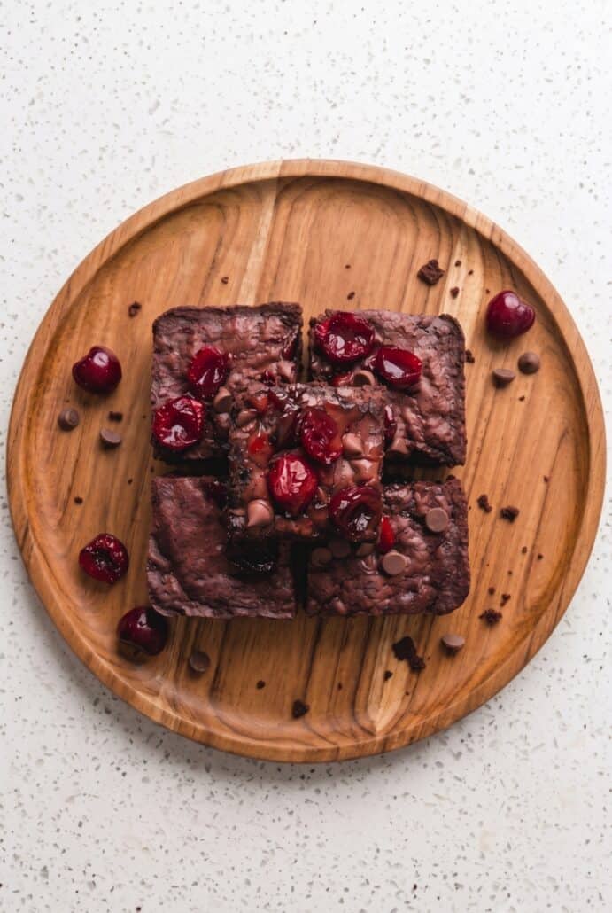 Stack of fudgy roasted cherry brownies topped with cherries and chocolate chips on a wooden plate.
