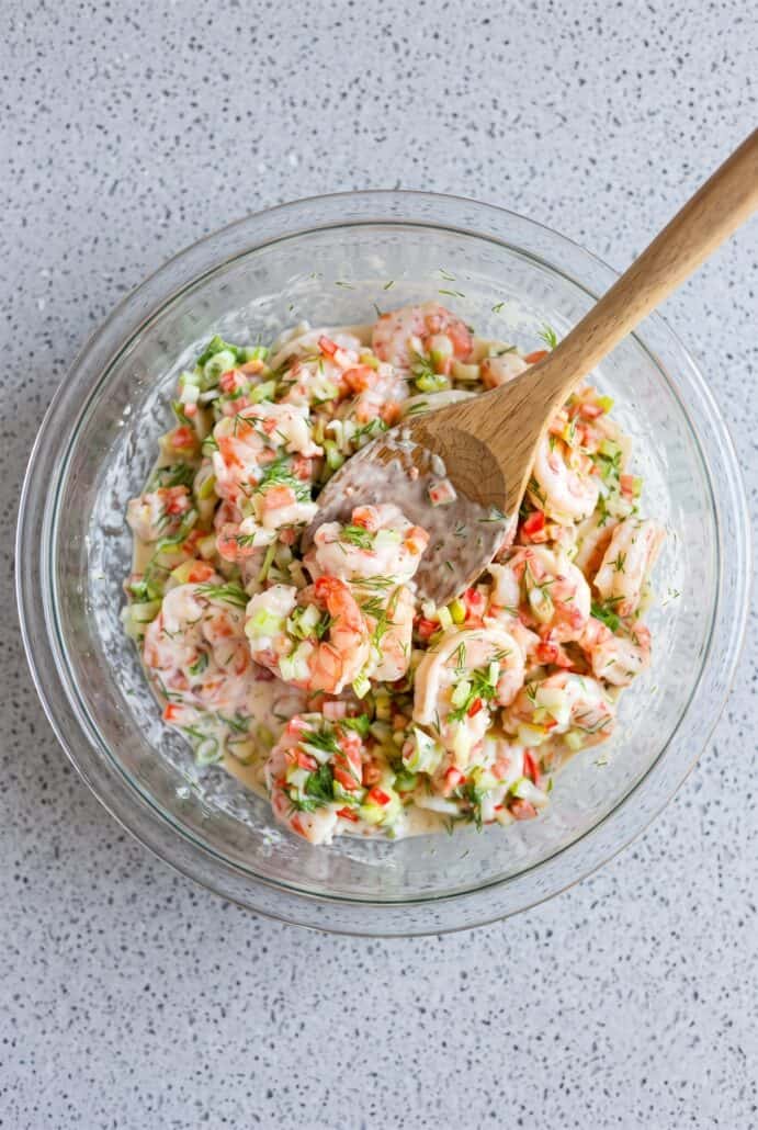 Mixing creamy shrimp salad with dill and chopped vegetables in a glass bowl.
