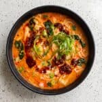 Bowl of spicy coconut noodles with creamy broth, chili oil, pak choi, scallions, cilantro, and lime.
