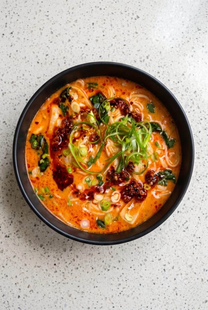 Bowl of spicy coconut noodles with creamy broth, chili oil, pak choi, scallions, cilantro, and lime.