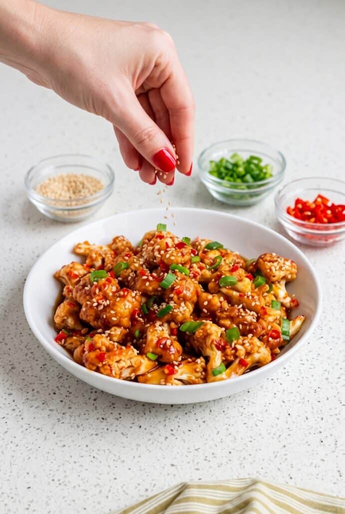 Hand sprinkling sesame seeds over sticky sesame cauliflower with green onions and red chilies for a flavorful vegetarian meal.