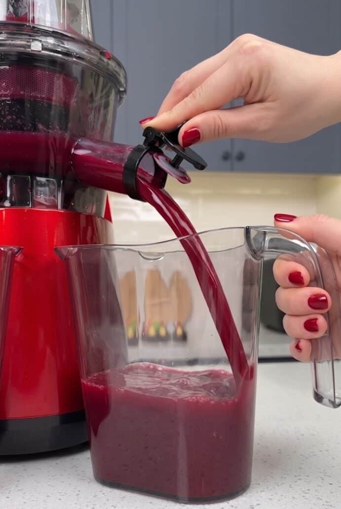 Fresh red fruit and vegetable juice pouring from a juicer into the collection pitcher.