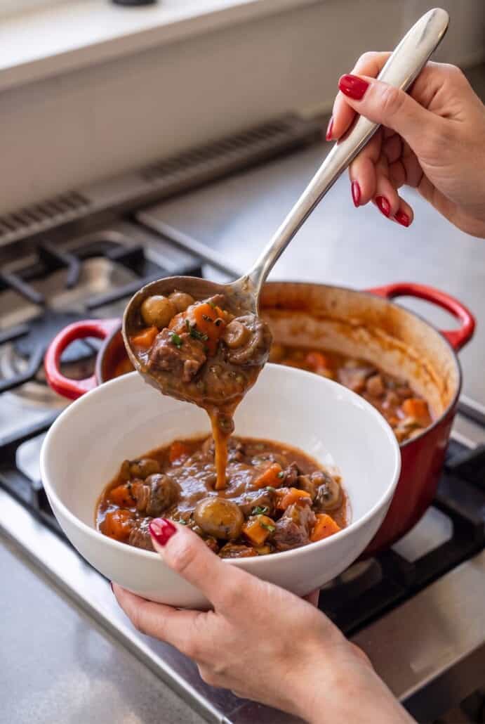 Ladling tender Beef Carbonnade Stew with carrots and mushrooms from a Dutch oven into a serving bowl.