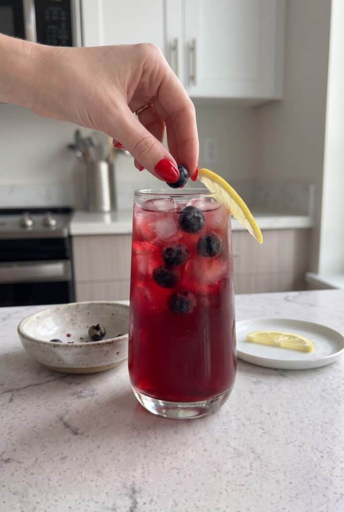 Hand adding a fresh blueberry to a glass of blueberry lemonade with lemon and ice.