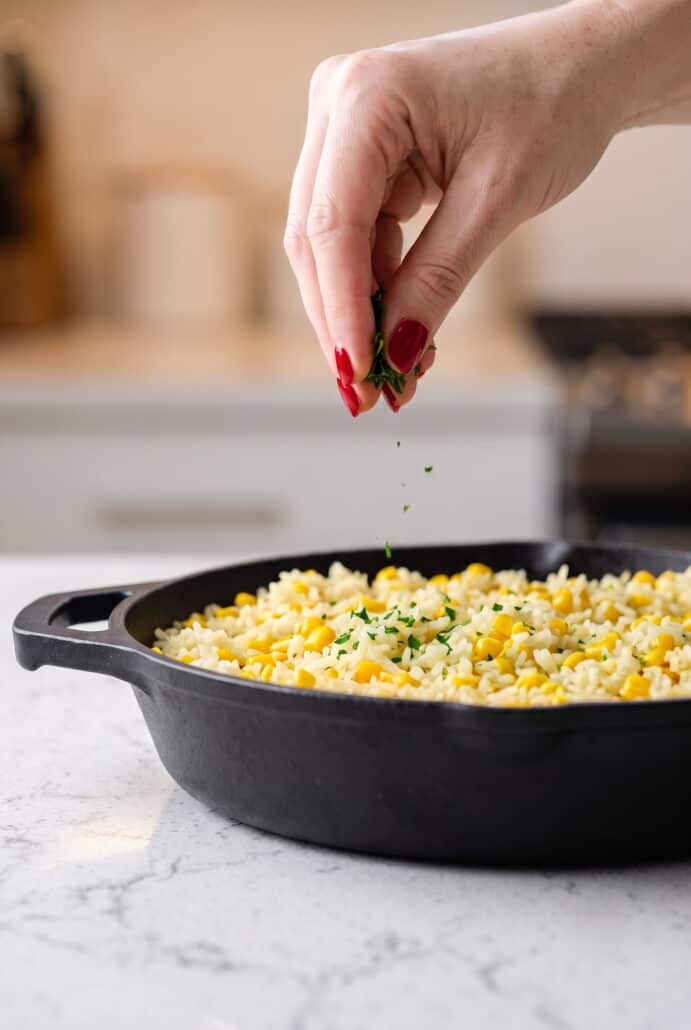 Fresh parsley being sprinkled over finished buttered corn rice in a skillet.