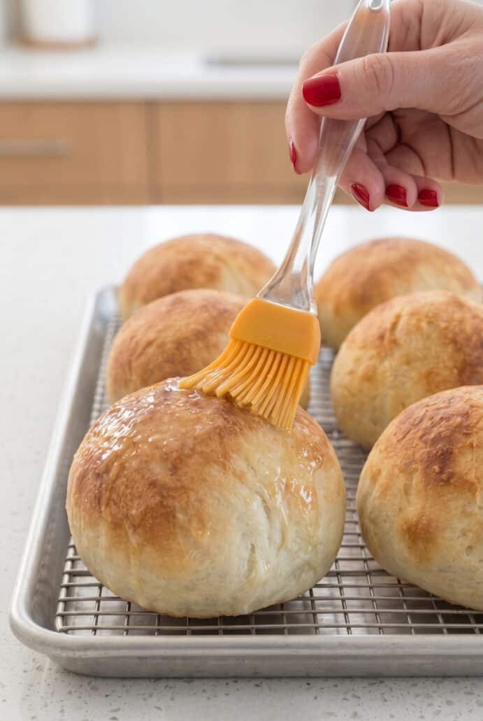 Brushing warm homemade bread bowls with butter after baking for a golden, flavorful crust.