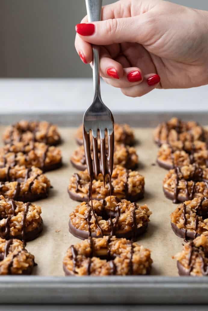 Fork drizzling melted chocolate over homemade Samoas cookies topped with caramel and toasted coconut.