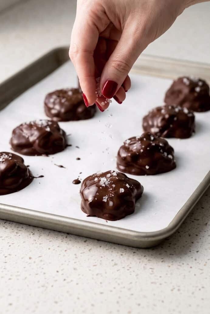 Hand sprinkling sea salt over chocolate-covered strawberry yogurt bites on a baking sheet.
