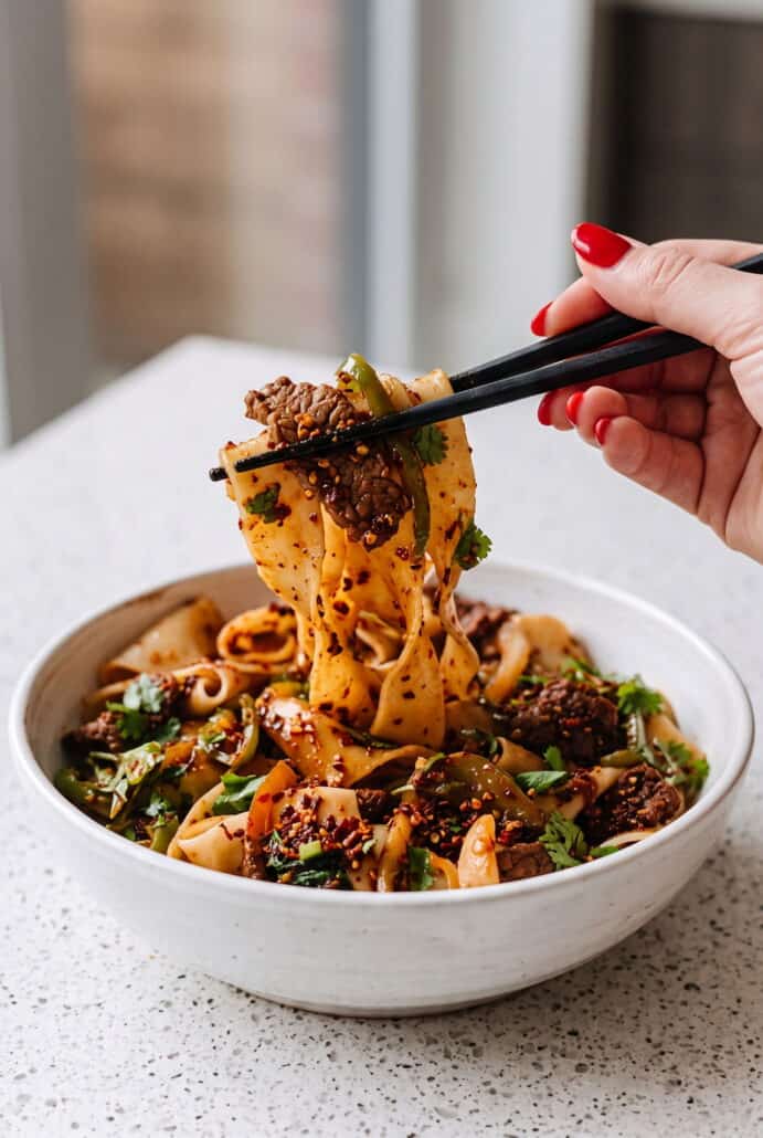 Chopsticks lifting spicy cumin lamb noodles with tender lamb and chili-seasoned wide noodles from a bowl.