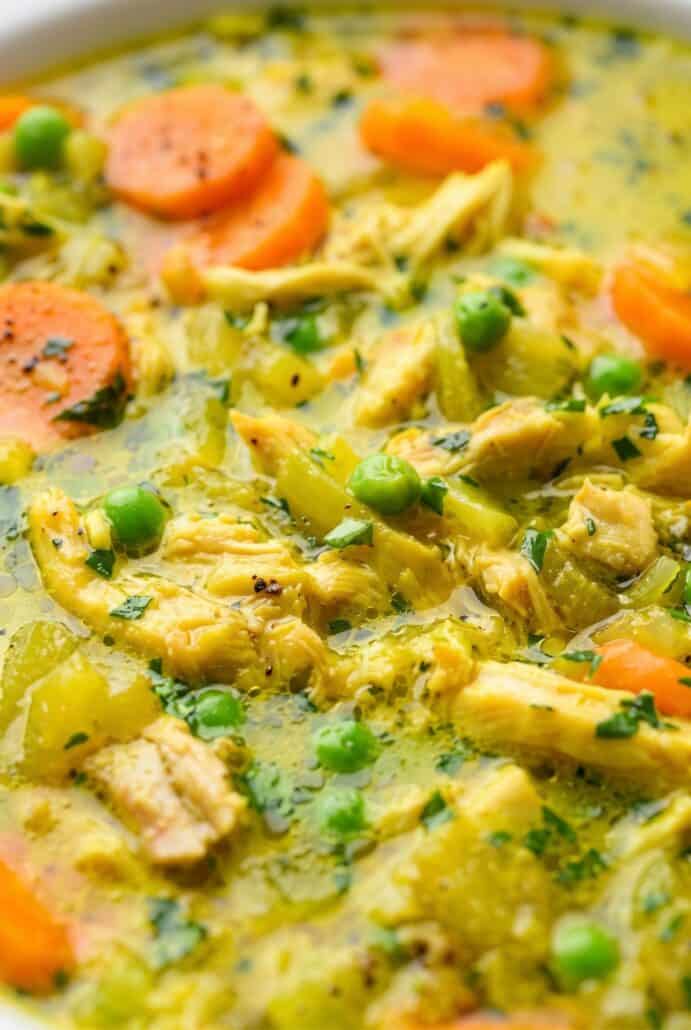 Close-up of turmeric chicken soup with tender chicken, carrots, peas, and herbs in a creamy golden broth.