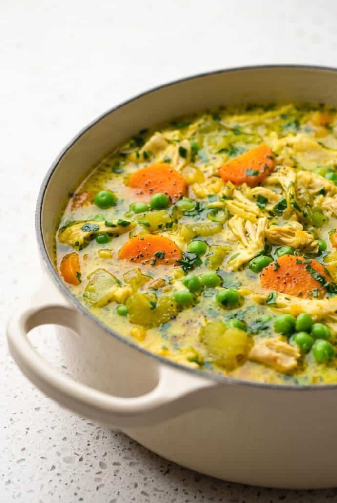 Close-up of turmeric chicken soup with shredded chicken, carrots, peas, and fresh herbs in a creamy golden broth.