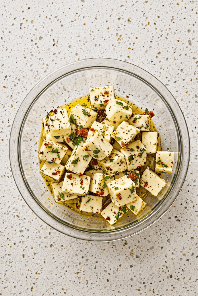 Bowl of marinated mozzarella cubes coated with herbs and red pepper flakes, ready to serve as an easy appetizer.