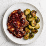 Raspberry balsamic glazed pork chop with roasted Brussels sprouts on a white plate.