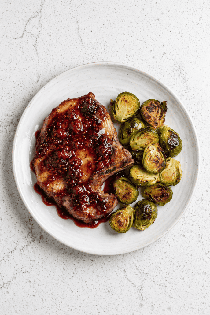 Raspberry balsamic glazed pork chop with roasted Brussels sprouts on a white plate.